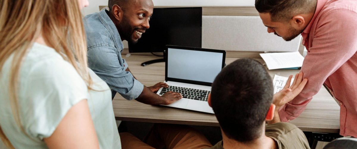Four colleagues brainstorming around a laptop in a bright, modern office setting.